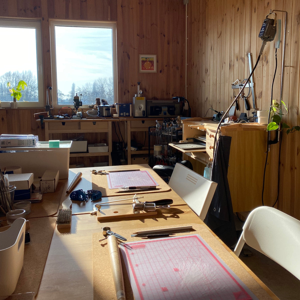 A cluttered but authentic wood workshop or craft studio with a long workbench in the foreground, tools, drawers, windows, and wood-paneled walls.