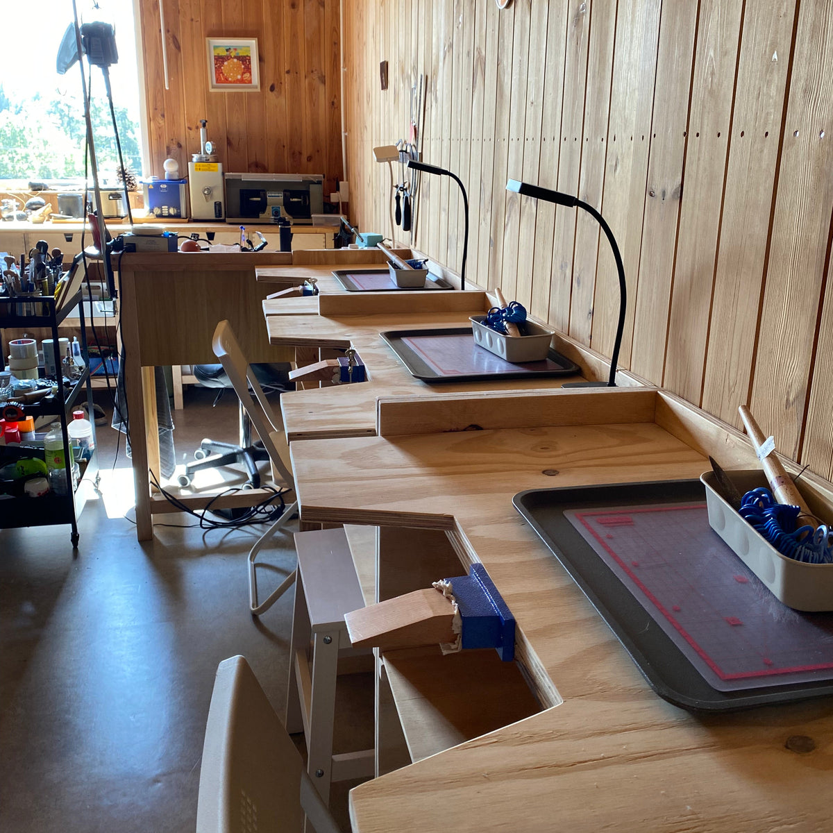 A workshop or craft studio with multiple light wood workbenches, chairs, desk lamps, trays, and tools arranged along a wood-paneled wall. No person is visible, but this is not a clean isolated product shot; it shows a real interior environment.